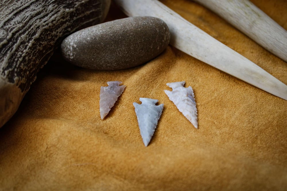 Three intricately knapped arrowheads made from flint or chert, displayed on a soft tan leather surface. Surrounding them are natural materials, including a smooth river stone, a large deer antler, and another piece of textured antler, emphasizing the traditional craftsmanship of flint knapping.