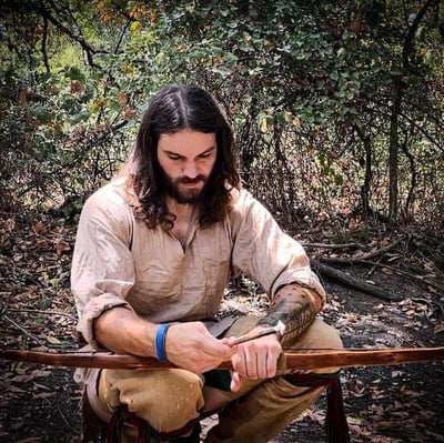 Phillip Liebel, an experienced flint knapper, sits outdoors in a wooded setting, with his handmade bow and arrow. Dressed in traditional-style clothing, he appears focused and immersed in his craft, surrounded by nature.