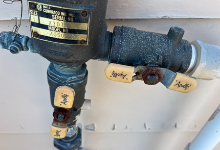 Close-up of an outdoor water shut-off valve system with rusted metal handles labeled 'Apollo' and 'good.' The system is mounted against a light-colored wall, showing signs of weathering and exposure.