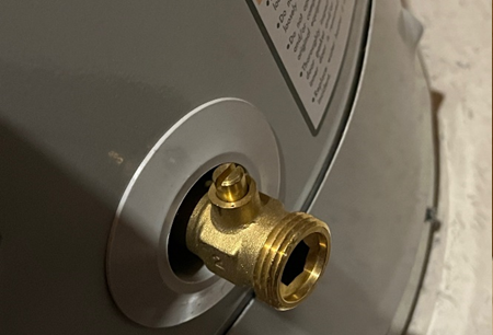 Close-up of a brass drain valve on the lower portion of a water heater, designed for maintenance and emergency water access. The threaded opening allows for hose attachment, facilitating controlled drainage.