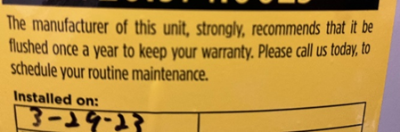 Close-up of a yellow maintenance label on a water heater, stating the manufacturer's recommendation to flush the unit annually to maintain the warranty. The installation date '3-19-23' is handwritten at the bottom.