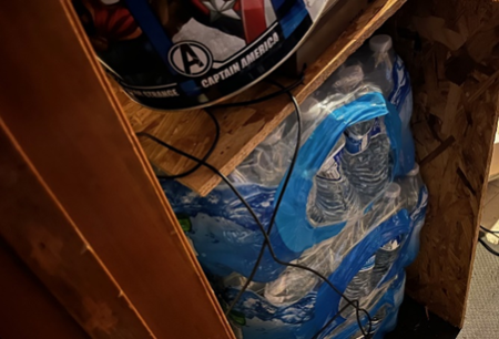 Stacked cases of bottled water stored in a small, enclosed space with wooden shelves above, featuring a Captain America-themed container and electrical cords running through the area.