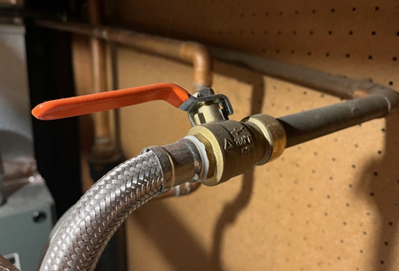 Close-up of a brass shut-off valve with an orange lever handle, connected to a flexible braided hose and copper piping, mounted against a pegboard wall in a utility area.