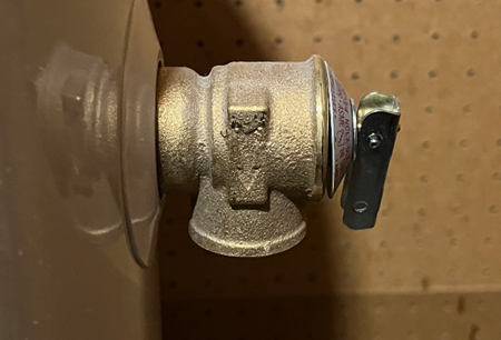 Close-up of a pressure relief valve on a water heater, featuring a brass body and a black lever for manual operation. Mounted against a pegboard wall, the valve is a key safety component designed to release excess pressure and prevent overheating.