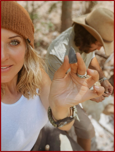 Jess Caldwell holds up a freshly knapped arrowhead with a smile, while Phillip Liebel works on another piece in the background. The outdoor setting and natural light highlight the hands-on flint knapping experience.