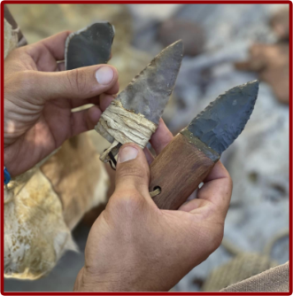 A pair of hands holding three handcrafted stone tools with flint or chert blades. One blade is wrapped with natural fiber cordage, while another is set into a wooden handle, showcasing traditional flint knapping and primitive tool-making techniques.