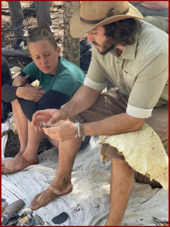 Phillip Liebel, wearing a cowboy hat and traditional-style clothing, sits barefoot on a blanket outdoors, demonstrating flint knapping techniques to a student. The ground is scattered with stone flakes and tools, creating a hands-on learning environment in a natural setting.