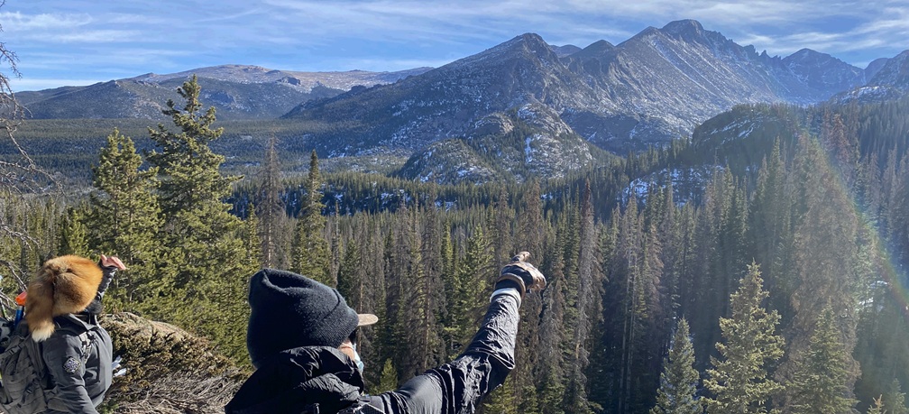 Two hikers overlooking a forested mountain valley, with one pointing toward the rocky peaks.