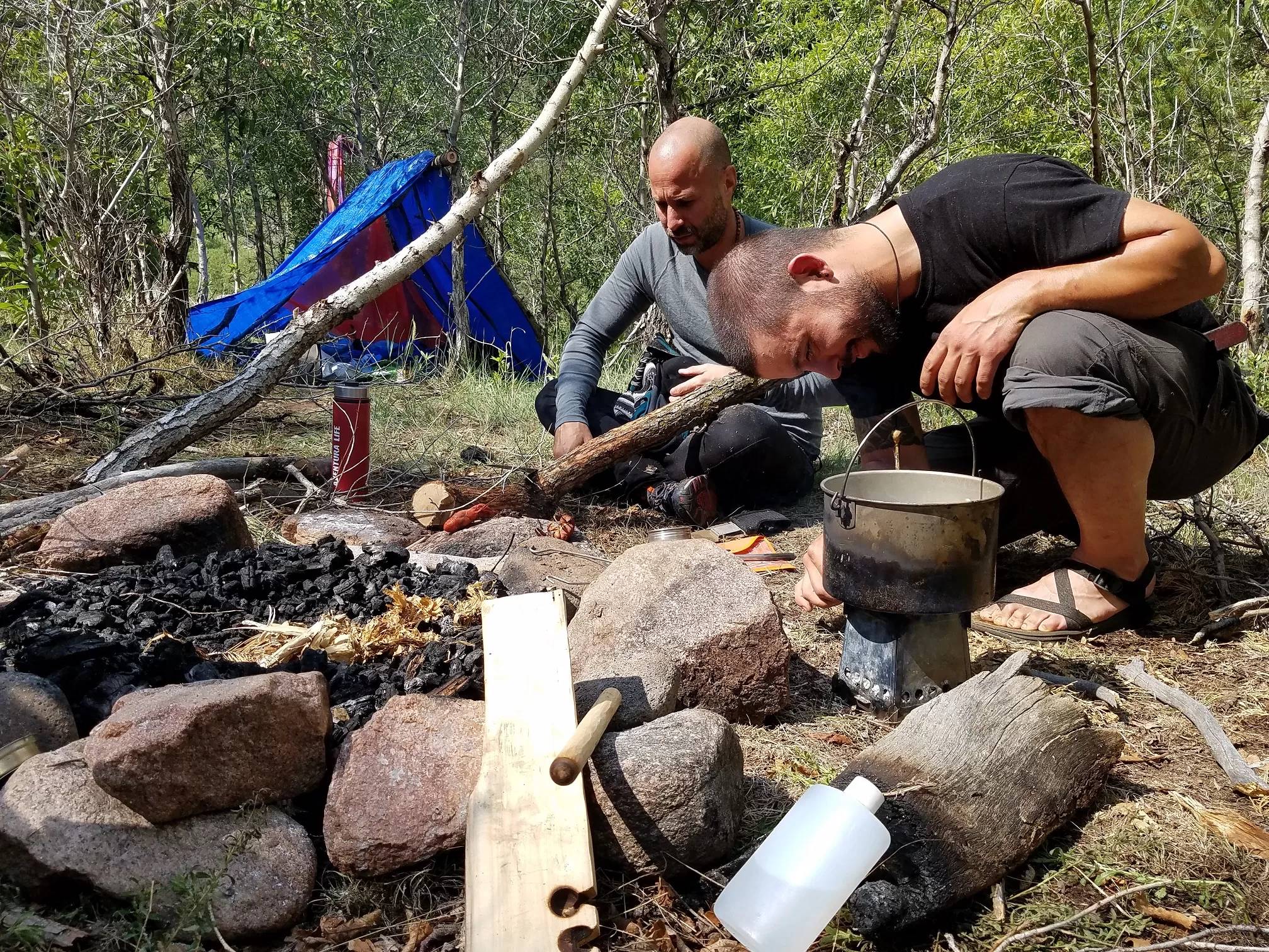Students practice campfire skills beside a tarp shelter during a beginner survival course.