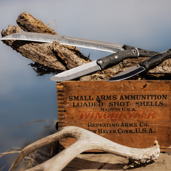Three fixed-blade knives from The Survival University’s Mountainside Mettle collection displayed on a vintage Winchester ammunition crate with driftwood and antlers, against a soft sky and natural backdrop.