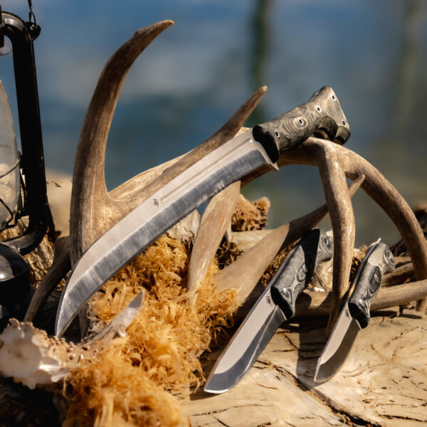 Display of The Spirit, The Courage, and Gallantry knives laid across antlers and wood shavings on a sunlit log, with a vintage lantern beside them. All knives feature micarta handles and rugged blades, set against a blurred lake background.