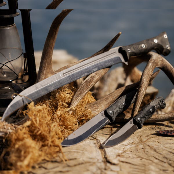 Three Mountainside Mettle knives displayed among antlers, curly wood shavings, and an obsidian arrowhead on a sunlit tree stump near water, evoking rugged outdoor craftsmanship.