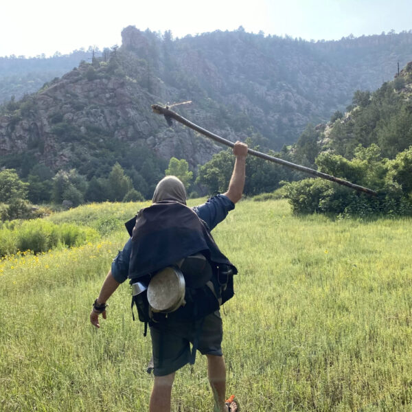 A person with a pot strapped to their backpack raises a long stick triumphantly while striding through a green mountain meadow toward rocky cliffs under a hazy sky.