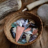 A woven basket filled with various colorful, hand-knapped arrowheads made of stone and glass. The basket rests on a fur hide, surrounded by traditional flintknapping tools including antlers and a large hammerstone.