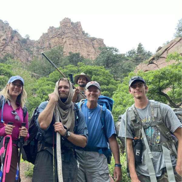 A group of six hikers smile for a photo at the base of a rugged red rock canyon. They're geared up with backpacks, trekking poles, and walking sticks, surrounded by green forest and steep cliffs.