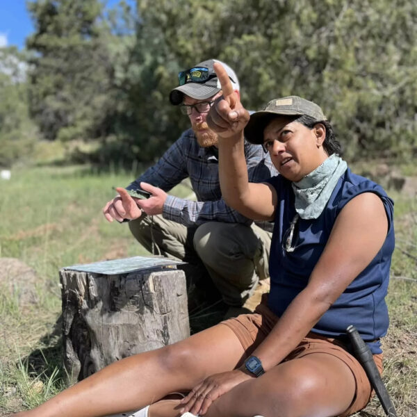 Two people sitting on the ground, one pointing while discussing navigation with a notebook and map nearby.