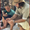 A barefoot instructor in a cowboy hat and students sit on a tarp in the forest during a flint knapping session, examining stone tools.