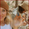 A woman in a white tank top and brown beanie holds up a small arrowhead toward the camera, smiling. A man in the background, wearing a tan hat, is focused on crafting or flint knapping.