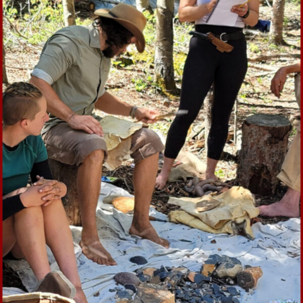 A barefoot man wearing a tan hat demonstrates flint knapping techniques on a tarp in the woods, surrounded by students and stone materials.