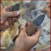 A close-up of hands holding three handmade flint blades, each mounted on different natural handles using sinew or plant cordage, showcasing primitive tool craftsmanship.