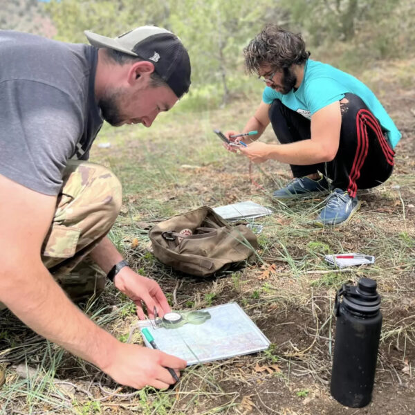 Two men crouch in a forest clearing, plotting bearings on topographic maps using compasses.