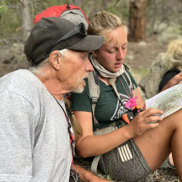 An older man and a younger woman sit in the forest studying a topographic map together, with compasses and outdoor gear visible, as they engage in a focused navigation lesson.