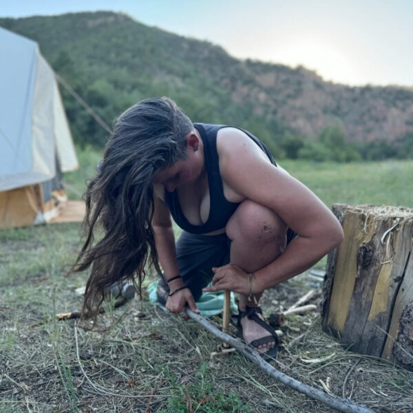 A woman with long hair, identified as Jacks, crouches on the ground working with a bow drill to start a fire, with a canvas tent and mountains in the background during golden hour.