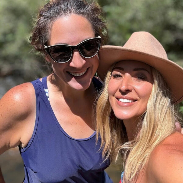 Two women pose for a sunny selfie during a river retreat. One wears a navy swimsuit and dark sunglasses with her hair up, while the other wears a tan sunhat and a red bikini top. The background is lush with green foliage and dappled sunlight.