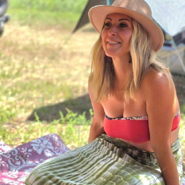 Jess sits outdoors on a patterned blanket under dappled sunlight, wearing a wide-brimmed hat, a red bikini top, and a green tie-dye skirt. Behind her, tents and gear suggest a riverside camping retreat.