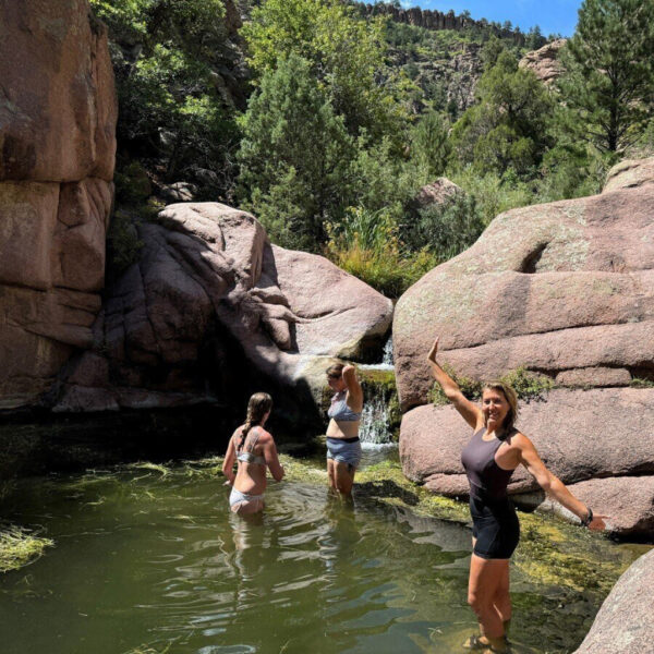 Jess smiles with arms outstretched in a natural swimming hole surrounded by red rock canyon walls, as two other women enjoy the water beneath a small waterfall.
