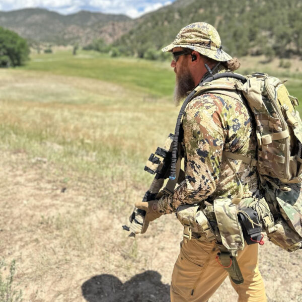 A man in camouflage gear and tactical backpack walks through an open mountain meadow under a bright blue sky, holding a rifle and wearing sunglasses and ear protection.