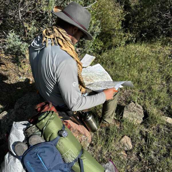 Man sits in tall grass studying a topographic map, surrounded by gear including a backpack, bedroll, and water bottle.