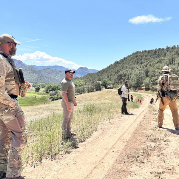 A group of individuals in tactical and outdoor gear are gathered on a dirt road in a scenic mountain valley, with some observing red flags placed in the ground, possibly during a field exercise or tracking course.