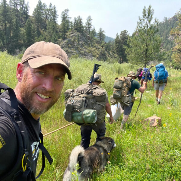 A smiling man in the foreground takes a selfie while leading a group of backpackers and a dog through a lush, green mountain meadow surrounded by trees and rocky hills.