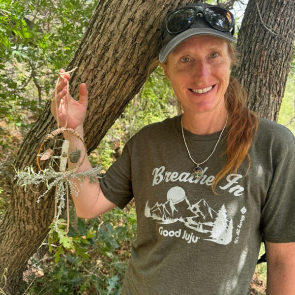 A woman wearing a forest green "Breathe In Good Juju" shirt and cap smiles while holding up a nature-crafted dreamcatcher made of twigs, leaves, and lichen, standing beside a large tree trunk in the woods.