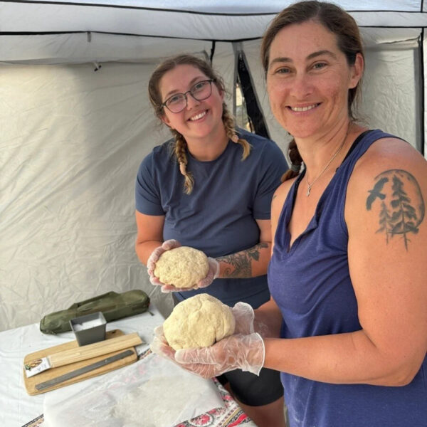 Two smiling women in a canopy tent hold balls of bread dough with gloved hands. A prep table in front of them is covered with parchment paper, measuring tools, and baking supplies.