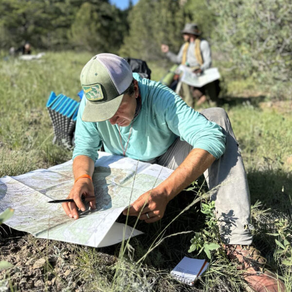 Man kneels in the grass, focused on plotting coordinates on a topographic map with a notebook nearby.