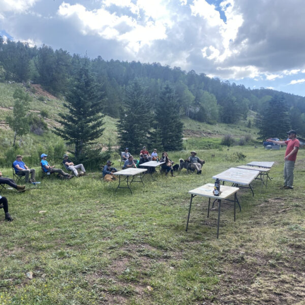 A group of people sit in chairs on a grassy hillside listening to an outdoor instructor near several folding tables and pine trees.