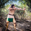 A man dressed in traditional buckskin attire is aiming a longbow in a wooded area, preparing to release an arrow. The forest floor is covered in leaves, and sunlight filters through the dense canopy above.