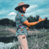 Phillip standing outdoors near a pond, wearing a wide-brimmed hat and a floral button-up shirt. He gestures with his right hand while looking forward, surrounded by dry grass and cloudy skies.