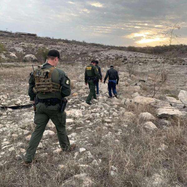 Law enforcement officers in tactical gear escort two individuals through a dry, rocky landscape at sunset.