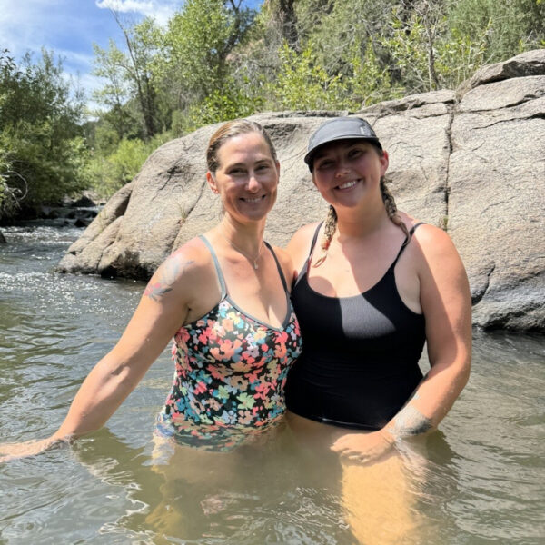 Two women smile while standing in a calm mountain river, waist-deep in the water. One wears a colorful floral swimsuit and the other a black swimsuit with a ballcap. They're surrounded by smooth boulders and lush greenery under a bright blue sky.
