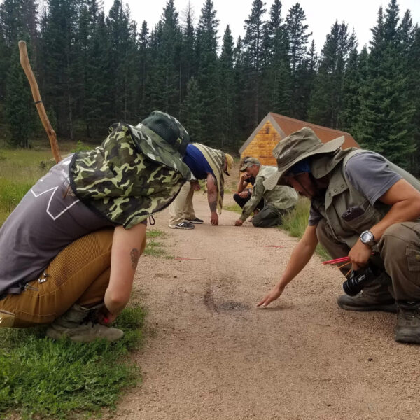 A group of people in outdoor gear crouch along a dirt trail inspecting sign during a tracking exercise, with a log cabin and pine forest in the background.