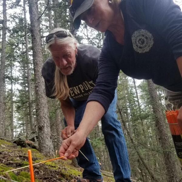 Two individuals practicing tracking skills in a forested environment, closely examining ground disturbances using orange tracking sticks during a Search and Rescue fundamentals course.