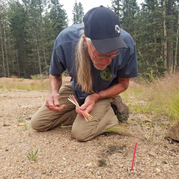 An older man with long hair and a black cap kneels on gravel, analyzing ground sign using colored tracking sticks during a SAR fundamentals course in a forest clearing.