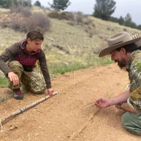 Two individuals kneel on a dirt path analyzing tracks. One uses a stick marked with blue string for measurements while the other places tracking sticks to mark impressions.