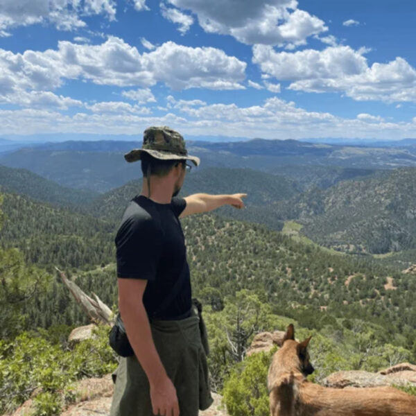 Man in camo hat points toward distant mountains while standing on a rocky overlook with a dog.