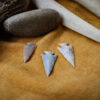 Three handcrafted stone arrowheads rest on a tan buckskin surface, surrounded by traditional flint knapping tools including a rounded stone and antler billets.