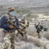 A tactical team moves uphill through arid, scrub-covered terrain. Members are in camouflage uniforms and gear, carrying rifles, with their faces obscured.