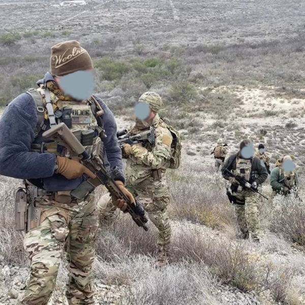 A tactical team moves uphill through arid, scrub-covered terrain. Members are in camouflage uniforms and gear, carrying rifles, with their faces obscured.